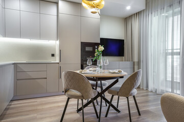 A contemporary dining area features a round table elegantly set with glassware and flowers, next to a sleek kitchen with modern appliances and soft lighting.