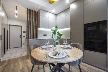 A well-designed modern kitchen features a round dining table set for two, with fresh flowers and stylish dinnerware in a contemporary apartment. The space is well-lit and inviting.