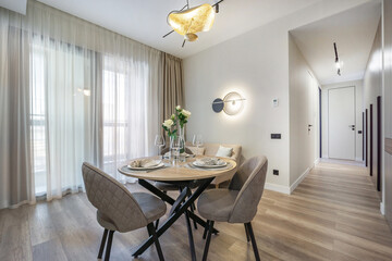 A stylish dining space features a round table set for two, surrounded by comfortable chairs, under a unique light fixture. The room is softly lit by sheer curtains.