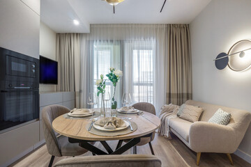 Cozy living space featuring a round wooden dining table set with elegant dishware. Comfortable sofa and tasteful decor enhance the inviting atmosphere. Natural light floods the area.