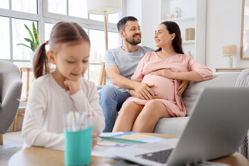 Happy pregnant couple sitting on sofa while their little daughter drawing at home