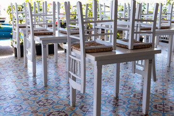 White wooden chairs stacked on tables in a closed Greek island restaurant, highlighting colorful tile flooring.