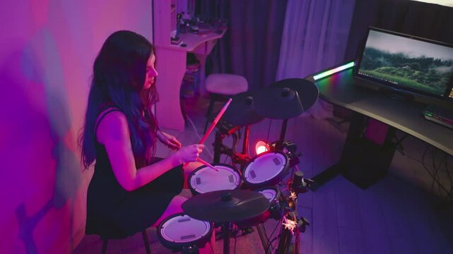 Young girl seated at electronic drum kit in studio lit with purple glow, playing with drumsticks while looking dull, monitor screen and desk setup visible nearby with musical equipment
