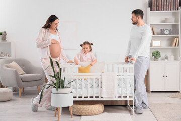 Cute little girl adjusting pillow on crib with her pregnant mother and father in bedroom