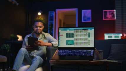Black remote worker navigating on tablet next to screen running analytics, sitting at home working on his daily tasks with multiple devices. Leisure and job productivity in loft. Camera A.