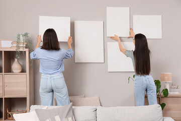 Young women hanging paintings on light wall at home, back view