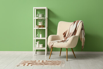 Stylish white armchair with soft plaid and shelving unit near green wall in room