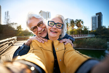 Cheerful retired couple taking a selfie, enjoying a sunny day in a city park, capturing memories together, embracing their golden years with smiles and laughter