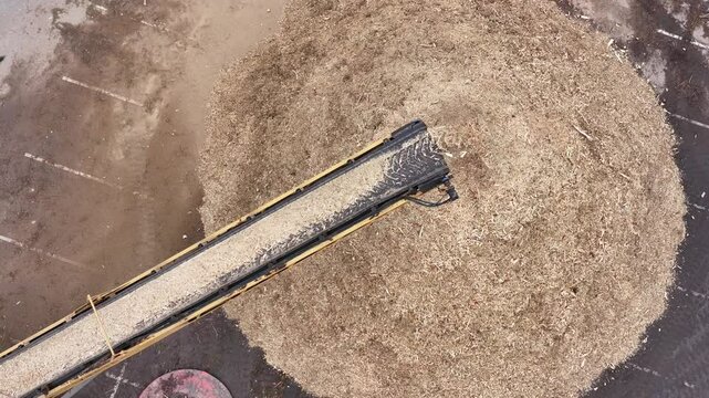 Slow-motion drone video captures a conveyor belt end discharging wood chips in a graceful cascade, each chip tumbling into a shifting pile below, set against the textured ground and warm, golden ambie