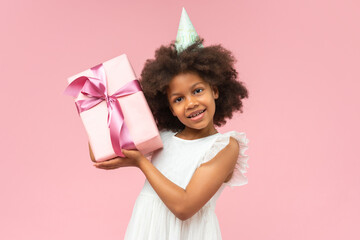 Adorable African American girl wearing white ruffle dress and birthday hat posing over pastel pink...