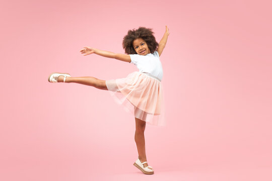 Full length studio portrait of adorable African American little girl showing classic dance step jumping and raising her leg, isolated over pastel pink background