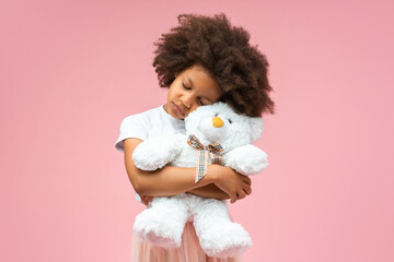 Studio portrait of adorable sleepy African American little girl hugging teddy bear toy, isolated over pastel pink background