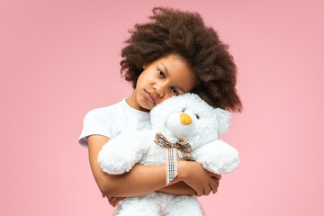 Close up studio portrait of cute African American little girl with sad face expression, hugging teddy bear trying to comfort herself, isolated over pastel pink background