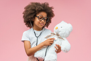 Studio shot of adorable African American girl wearing toy goggles playing doctor using stethoscope to check respiratory system of teddy bear