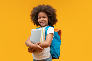 Studio portrait of joyful African American schoolgirl posing in studio with her backpack holding digital tablet and pile of books, isolated on bright colored orange yellow background