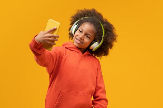 Studio portrait of cute confident African American little blogger girl listening to music via wireless headphones and taking selfie on her mobile phone