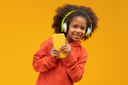 Studio portrait of cute smiling African American girl wearing wireless headset and posing with mobile phone in hands, isolated over bright colored orange yellow background
