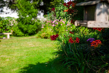 Beautiful backyard green garden with colorful flowers and fresh, green lawn background with a lot of space. Out of focus bird feeder in the foreground.