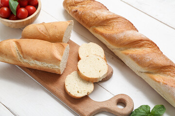 Board with cut fresh baguette, basil and cherry tomatoes on white wooden background, closeup