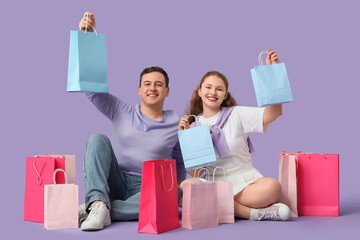 Beautiful young couple with shopping bags sitting on lilac background