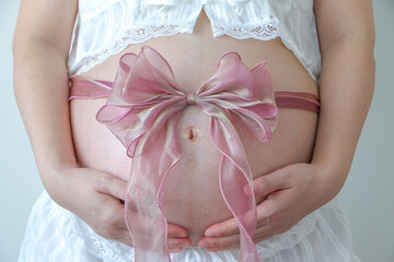 Close up of young pregnant woman’s hands holding her belly baby bump, wrapped in pink ribbon, symbol of precious gift of life. Pregnancy, maternity and expectation concept.