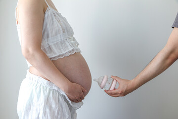 Expectant mother holds hands beneath her pregnant belly as father-to-be holds baby bottle towards the beautiful baby bump. Pregnancy, feeding baby, parenthood, expectation concept.