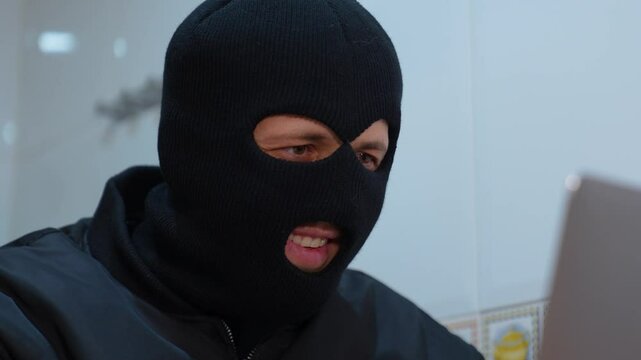 Hacker wearing balaclava is surprised while working on his laptop