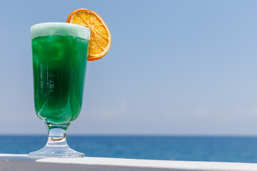 Refreshing green non-alcoholic cocktail with orange slice in glass with backdrop of blue sea. Perfect for summer relaxation and tropical getaways, travel holiday concept. Copy space.