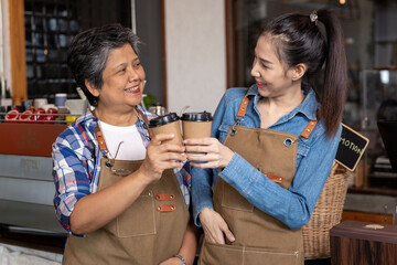 Asian middle-aged woman and young woman smiling while clinking takeaway coffee cups. Both wear brown aprons, casual shirts, stand in cozy coffee shop. Friendly cheerful moment shows happiness
