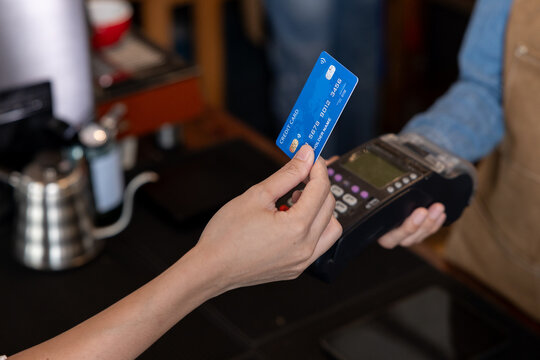 Close-up of customer hand holding blue credit card making electronic payment using point of sale terminal held by barista in small coffee shop representing modern contactless transaction service - Powered by Adobe