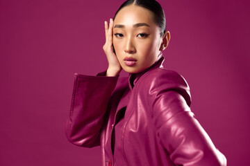 Fashionable asian woman in purple leather jacket posing with eyes closed and hand on head on purple...