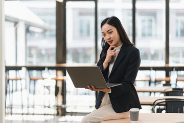 Fototapeta premium Young Asian businesswoman in black suit holding laptop and thinking in modern office, natural light, professional and focused atmosphere, coffee cup on table