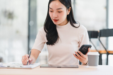 Young Asian woman working at desk with smartphone, tablet, and coffee cup, writing notes on paper...
