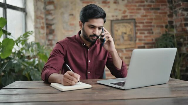 Man working at laptop, taking notes, talking on phone, modern office