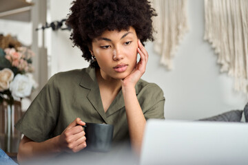 Cool gen z African American girl using laptop sitting at home drinking coffee. Young pretty Afro woman looking at computer relaxing with cup of tea in hands watching online tv series or movie.