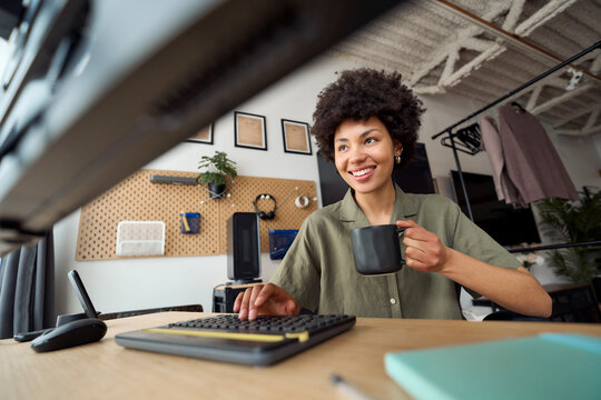 Young happy African woman student using pc typing elearning or hybrid working at home looking at computer learning, studying online, drinking coffee sitting at work desk. Low angle candid shot.