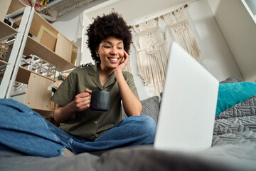 Happy gen z African American girl using laptop sitting at home bed drinking coffee. Young smiling...