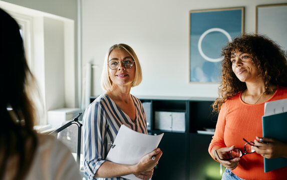 Smiling woman with blonde hair in a striped shirt is holding papers, likely presenting or preparing for a meeting. With her is a colleague in an orange top. They talk together.