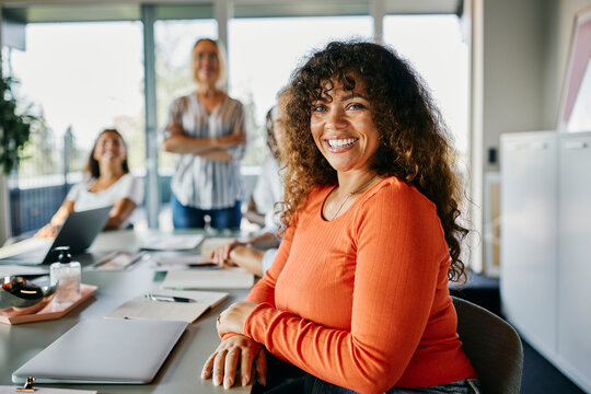 Confident woman with curly hair and orange top smiling in modern office setting. Documents, laptop, and snacks are spread out on the meeting table. Bright and positive atmosphere - Powered by Adobe