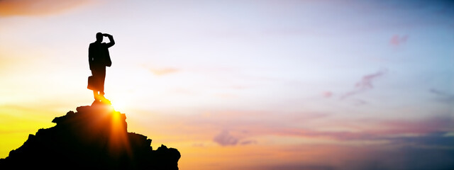 Businessman standing on mountain peak at sunrise looking forward with hand over eyes, symbolizing...
