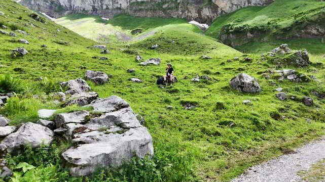 9 juli 2024, Wildhaus, Switzerland. Drone flight alpine meadows and hiking trail. A caucasian hiker with hat is sitting on a rock in the grass eating. Wonderful outdoors adventure. Great hiking spot, 