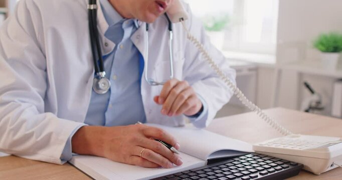 Portrait of a confident young male doctor working at his workplace with pc computer at office and talking by phone consulting sick patients in medical clinic. Medicine concept. 4k video
