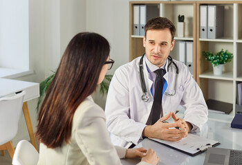 Young confident friendly male doctor consulting his woman patient sitting on workplace in...