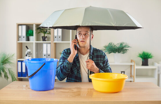 Worried man sitting at desk under umbrella, making phone call to repair and maintenance service. Office worker dealing with plumbing or roof problem and leaking water dripping into bucket and basin.
