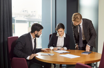 Business team discussing contract at round table in office. Smiling businesswoman signing agreement while two male colleagues in suits assisting, reviewing documents and sharing professional advice.