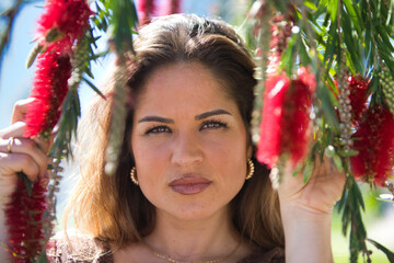 Portrait of beautiful young blonde woman among the red flowers hanging from a tree in the park. The woman is serene and with bright eyes.