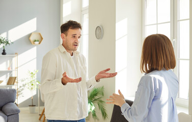 Frustrated young family couple standing by window and arguing with each other. Husband and wife expressing anger and disappointment, struggling with relationship issues and lack of understanding.