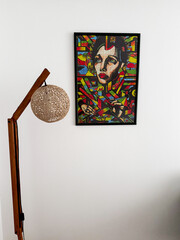 Minimal interior design featuring a modern wooden floor lamp and colorful abstract portrait art on a white wall. Ideal for concepts related to contemporary home decor and creative living spaces.