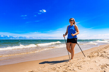 Playing sports on beach in summer. Attractive middle-aged woman practicing Nordic walking on sandy...