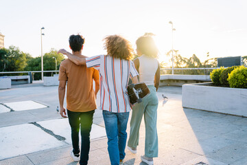 Three young friends walking away from camera at sunset in urban rooftop with skateboard, enjoying...